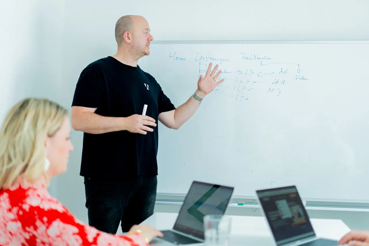 Frank Ewerle skizziert eine Seitenstruktur am Whiteboard, Kollegen verfolgen es am Laptop